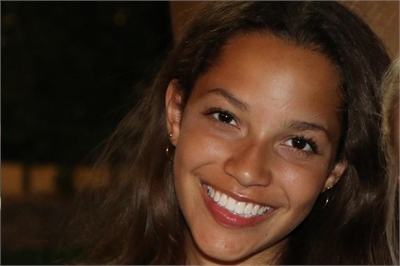 [ai] A smiling young woman with long, wavy hair and a warm expression, standing in a dimly lit outdoor setting. She is wearing simple earrings, suggesting a casual atmosphere.